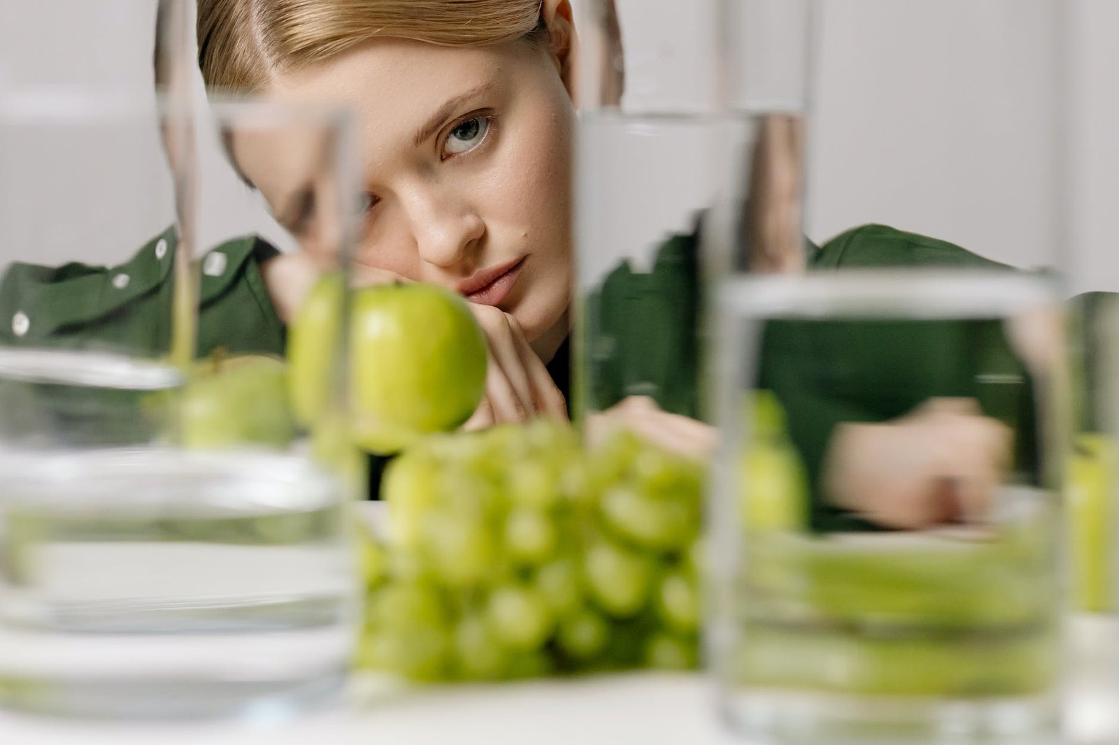 woman in green long sleeves near green fruits and glasses of water while seriously looking at the camera
